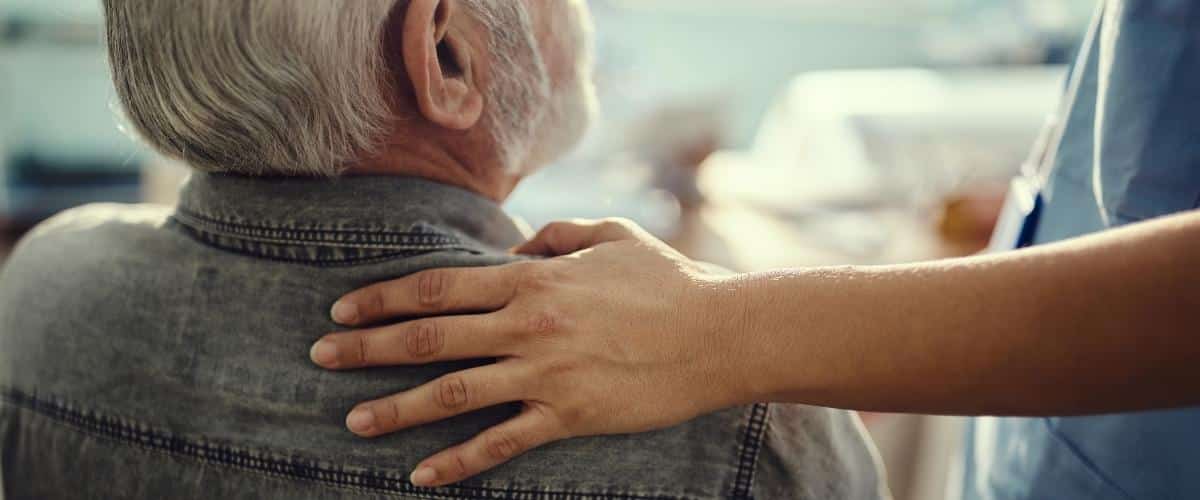 Eine Pflegerin legt tröstend die Hand auf die Schulter eines älteren Herren.
