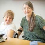 Begleitperson unterstützt eine sitzende Person bei einer Tätigkeit am Tisch, beide grinsen in die Kamera.