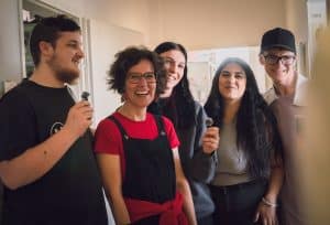 Ein Gruppenfoto von den Bigsteilnehmer*innen mit der Ausbildnerin. Zwei Personen halten Mikrofone und wirken wie bei einer Präsentation.