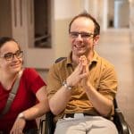 Zwei Personen sind nebeneinander, eine davon im Rollstuhl die andere am Boden hockend, im Gespräch.