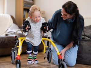 Eine Mutter begleitet ihren Sohn der mit einer Gehhilfe gehen lernt.