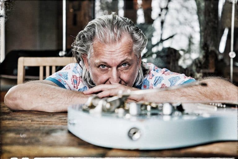 Sir Oliver Mally sieht über seine Gitarre während er sich auf den Tisch stützt.